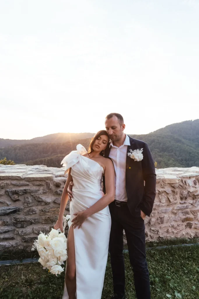 A couple at their Elopement in Medvedgrad, Zagreb, standing close to each other with a scenic view of mountains and sunset in the background.