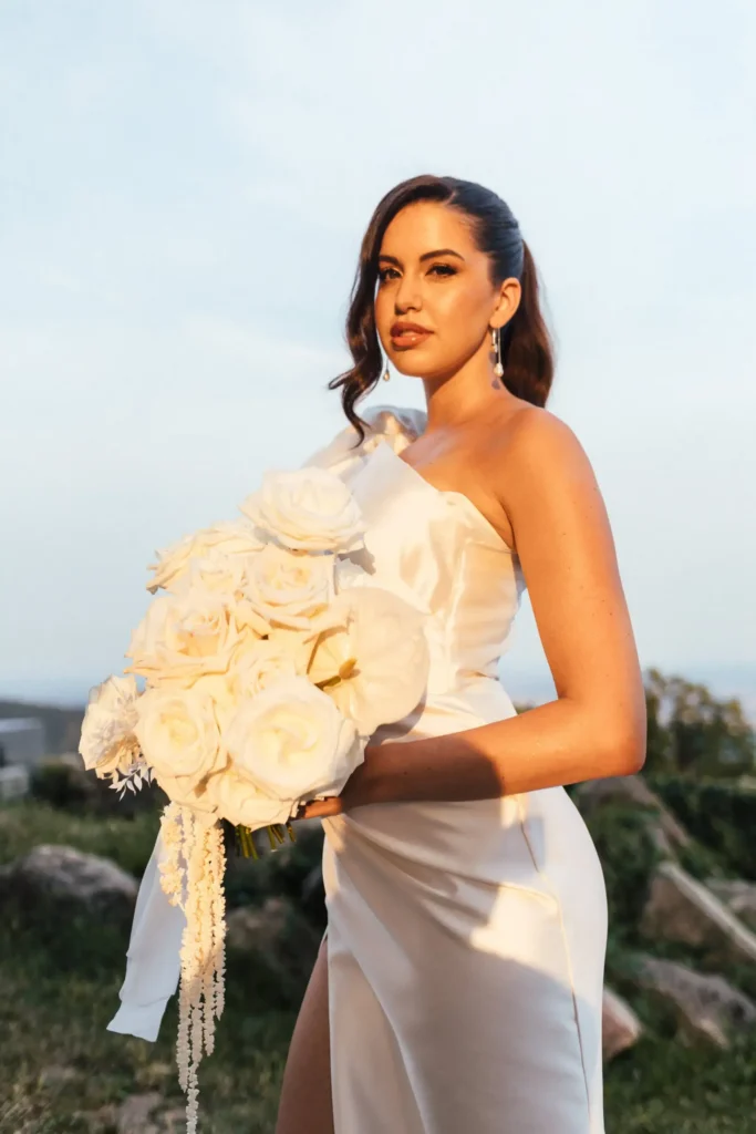 A person holding a bouquet of white roses and wearing an elegant white dress, standing outdoors with a scenic view in the background.