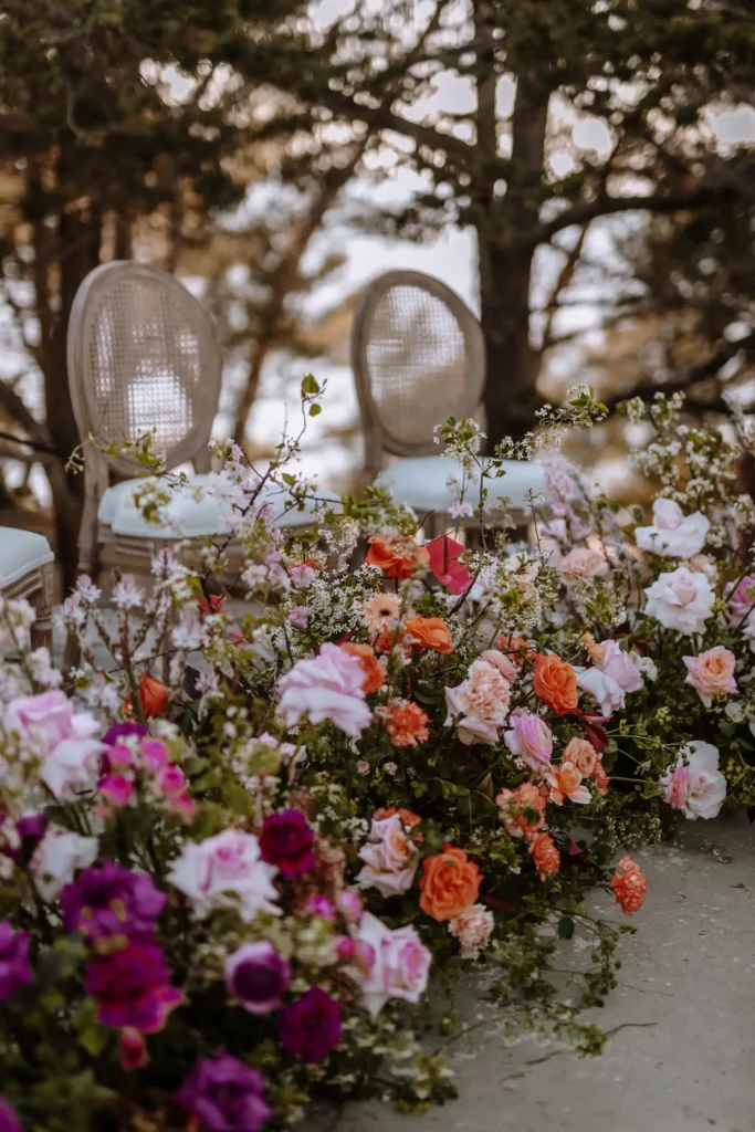 A colorful array of flowers in full bloom, including roses and petunias, set before two elegant wicker chairs amidst a serene outdoor setting.