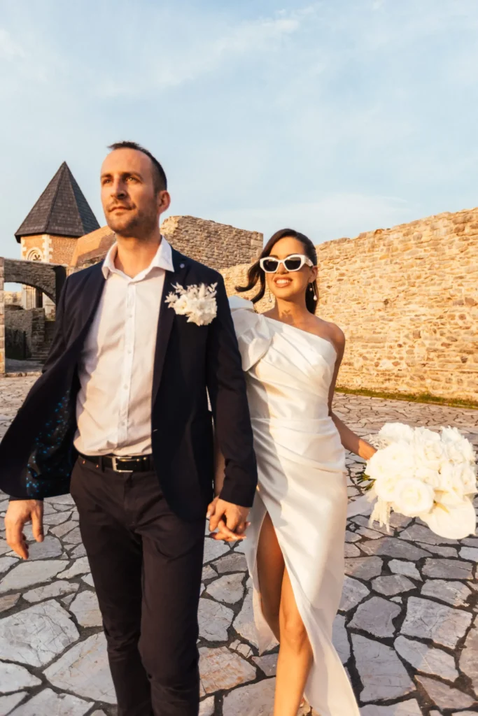 Bride in a white dress with a large bouquet and white-framed sunglasses walking hand-in-hand with her partner against the backdrop of Medvedgrad’s historic walls and tower.