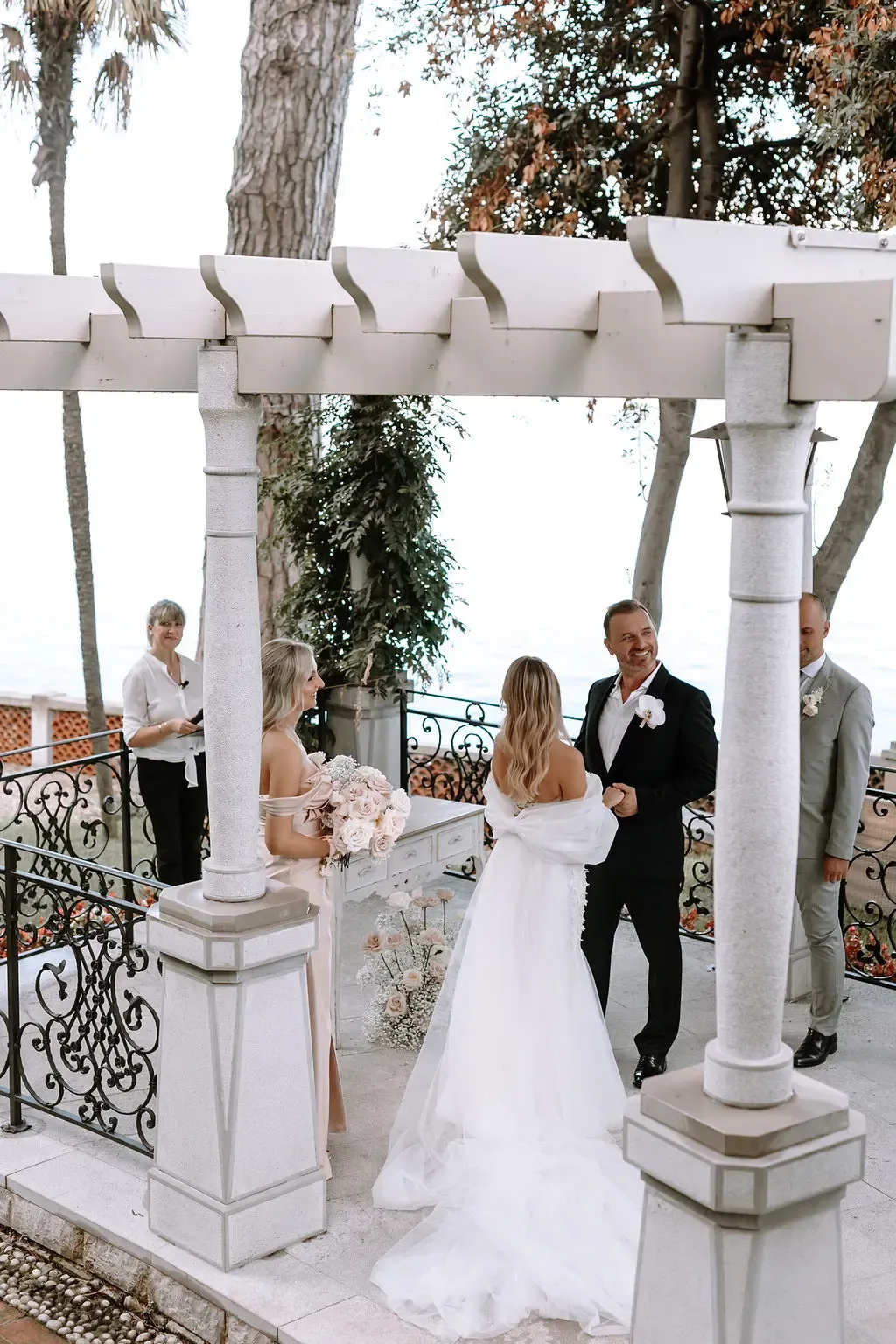 A couple exchanging vows at an outdoor wedding ceremony, surrounded by a few guests, with a scenic view of trees and the sky in the background.