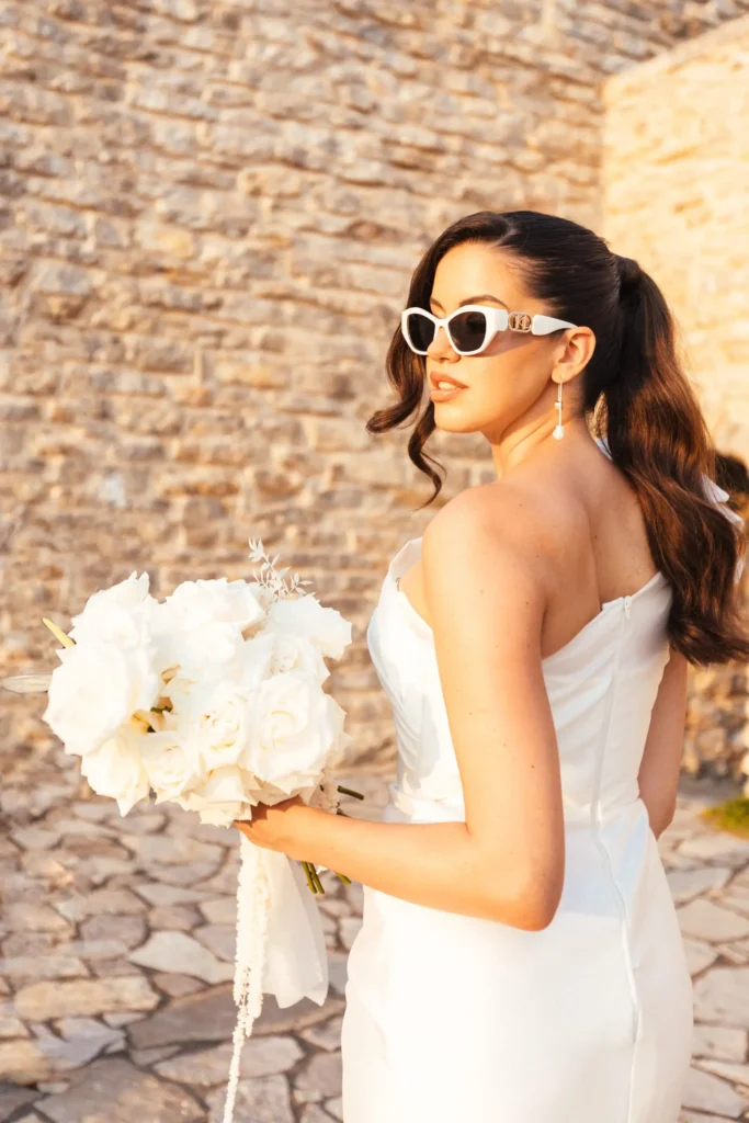 A bride in a stunning white dress holds a bouquet of white flowers, standing against a rustic stone wall.