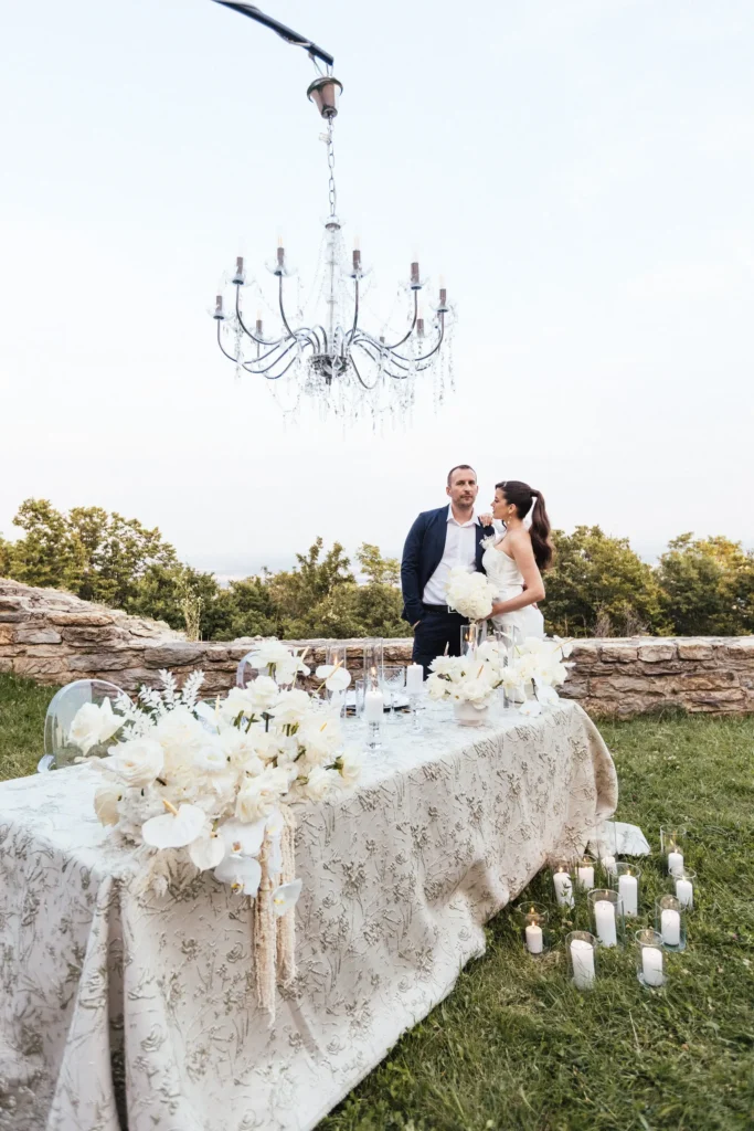 A couple holding hands and smiling, enjoying a romantic elopement dinner at a table decorated with candles and white flowers, under the starry sky and the majestic Medvedgrad Castle in Zagreb.
