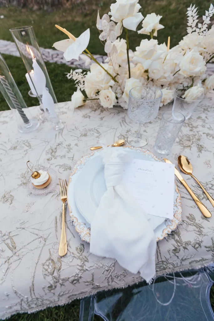 A beautifully decorated table setting featuring golden cutlery, white plates, a neatly folded napkin, and an exquisite white floral arrangement.