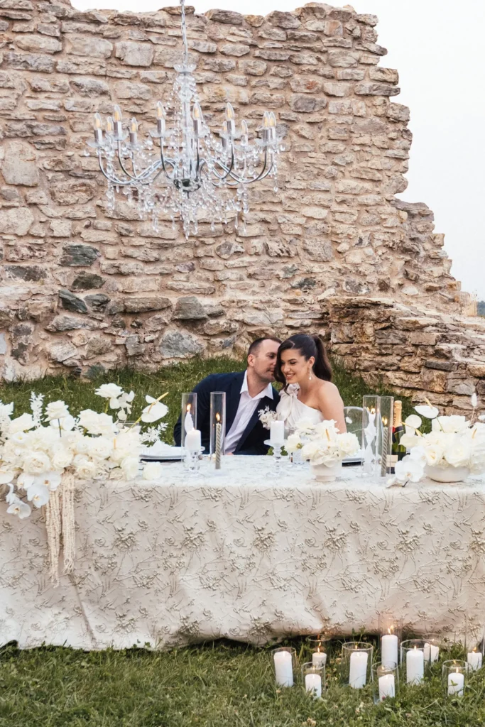 A couple holding hands and smiling, enjoying a romantic elopement dinner at a table decorated with candles and white flowers, under the starry sky and the majestic Medvedgrad Castle in Zagreb.