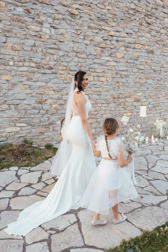 A bride in a white gown and a flower girl in a white dress walking hand-in-hand against a stone wall backdrop.