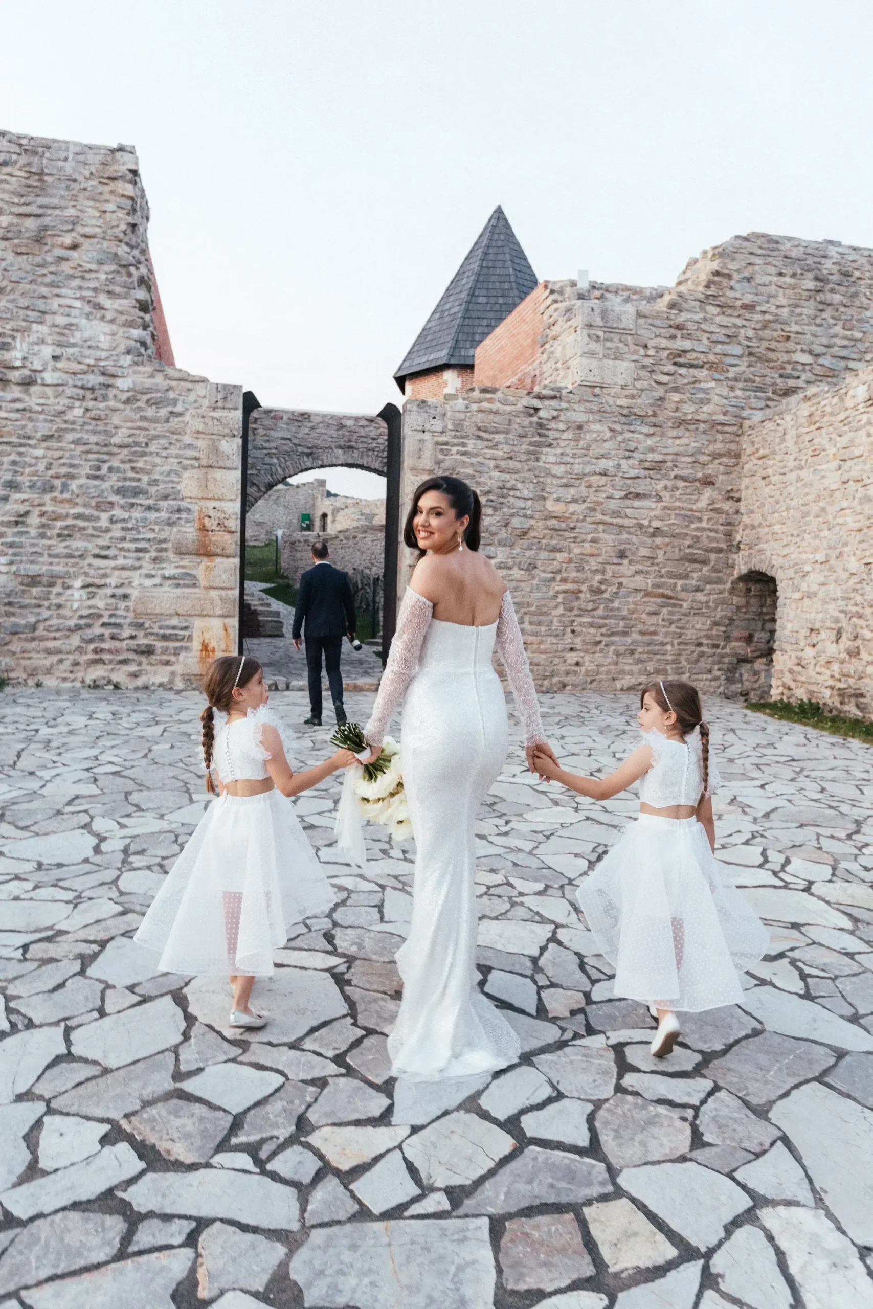 A person in a wedding dress holding hands with two children in white dresses, walking towards another person near an ancient Medvedgrad stone structure.