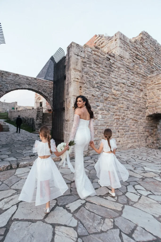 A bride in a white dress holding hands with two flower girls in front of an ancient stone castle during the golden hour.