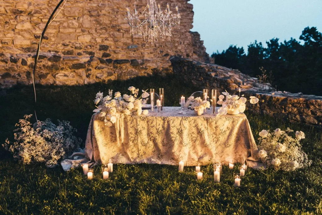 An elegantly set dining table adorned with flowers and candles, positioned outdoors under a crystal chandelier, with an ancient stone wall in the background.