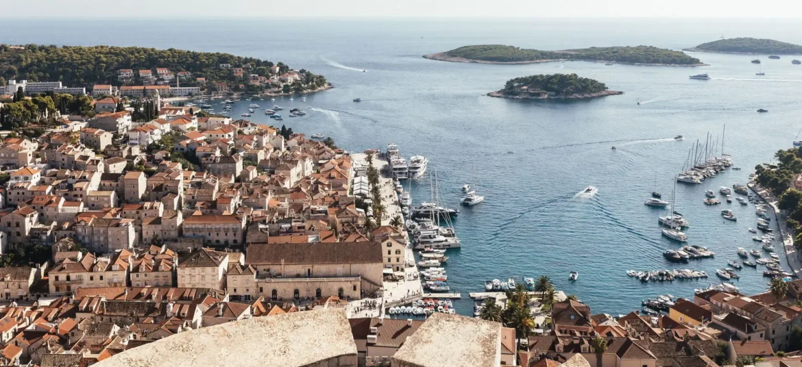A panoramic view of a coastal town with terracotta rooftops, a marina filled with boats, and surrounding islands, as seen from Hvar Fortress.