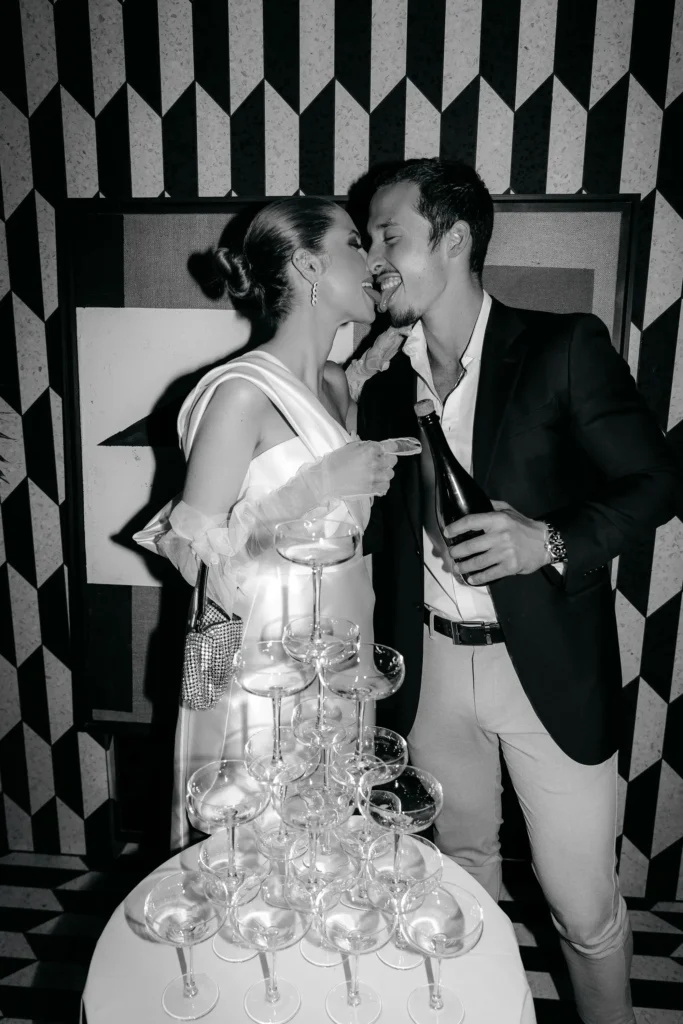 Two people standing beside a champagne tower, with their faces obscured, in front of a geometric patterned wall.