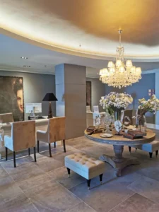 An elegant lobby area featuring a crystal chandelier, a round table adorned with flowers and decor, plush seating, and a large portrait on the wall.