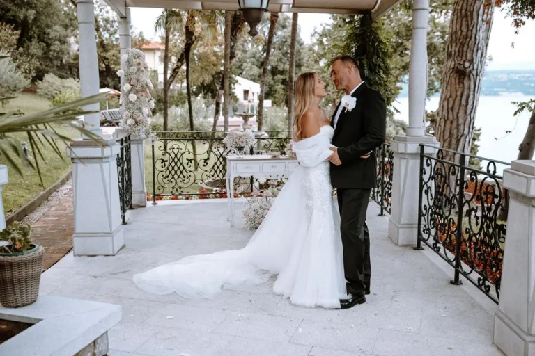 A couple in wedding attire embracing each other under a gazebo, surrounded by lush greenery and a serene sea in the background.